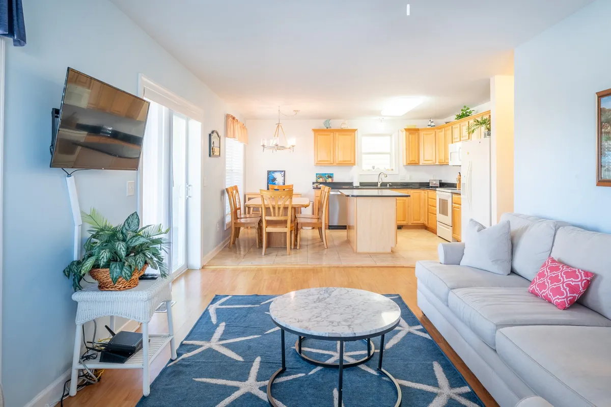 A wide shot capturing the open-concept living, dining, and kitchen area. The image features a plush sofa and coffee table in the living area, a dining table with enough seating for six, and a kitchen with modern appliances and visible cabinetry. The space is bright, cheerful, and designed for social gatherings, with the television also in view.