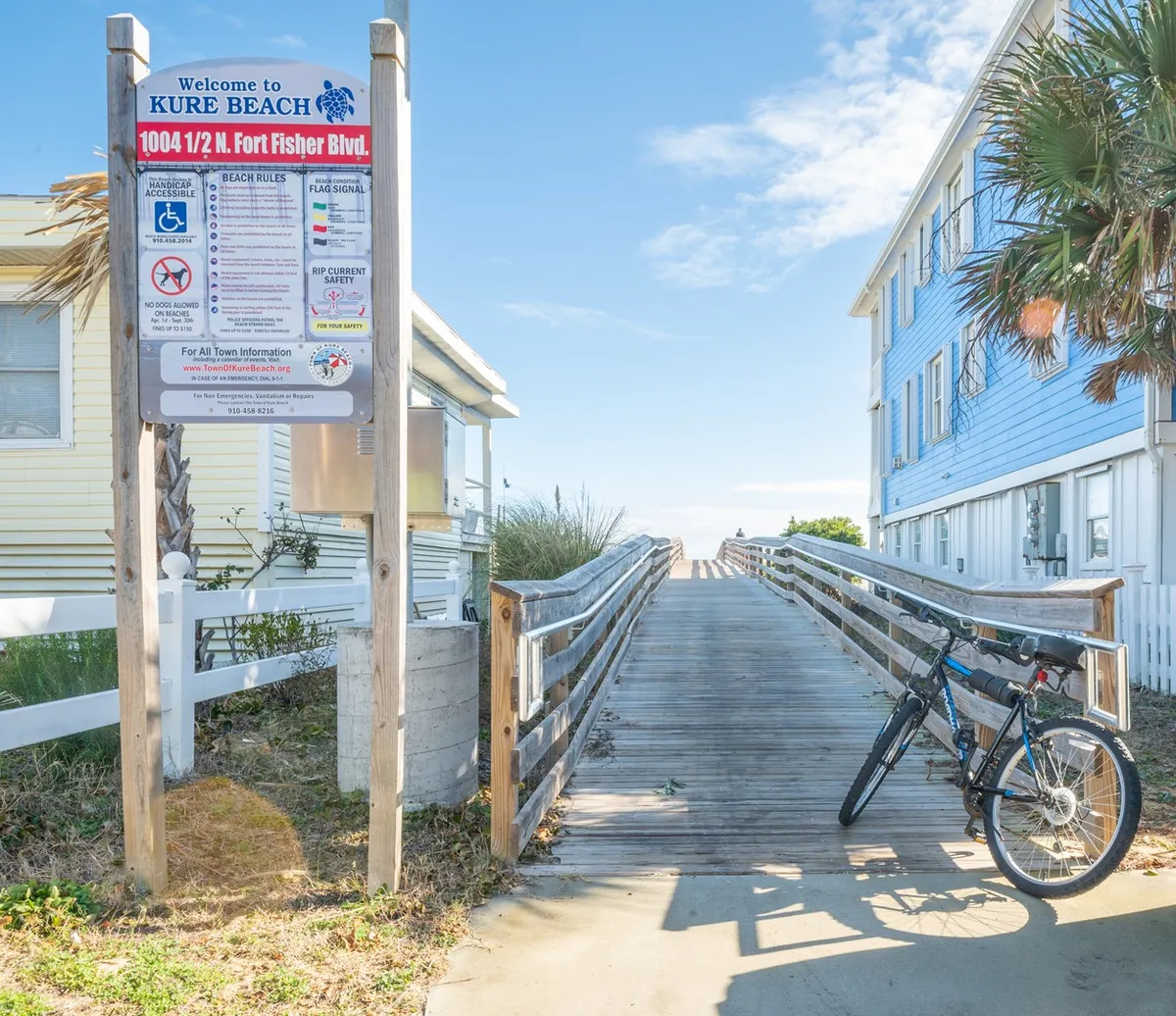 An inviting exterior image of the condo's entry area, with a welcoming sign, a parked bicycle, and a wooden walkway leading toward the property. The scene conveys the peaceful arrival experience and connection to the coastal neighborhood.