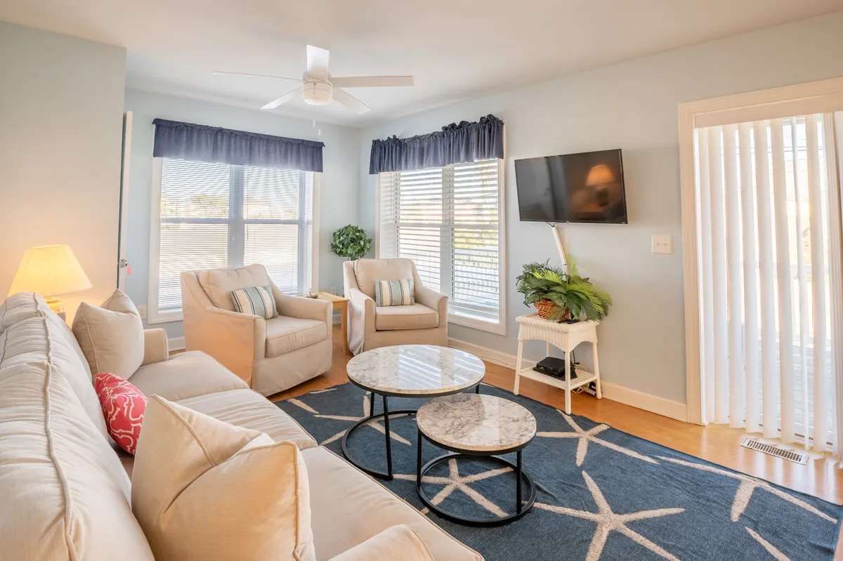 A cozy living room scene with a sofa adorned with decorative pillows, armchairs, a coffee table, a television, and a window with blinds letting in natural light. The arrangement invites relaxation, movie nights, and socializing in a comfortable, homey setting.