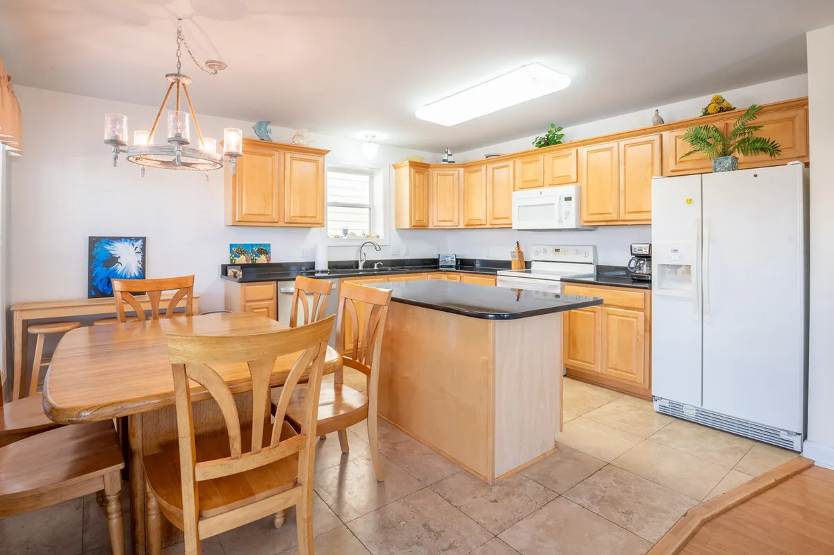 The image showcases the kitchen island and adjacent dining table, surrounded by wooden cabinets and white appliances. There is a sense of activity, as if a meal is about to be prepared or enjoyed, with seating for a group to gather around the table or kitchen island.