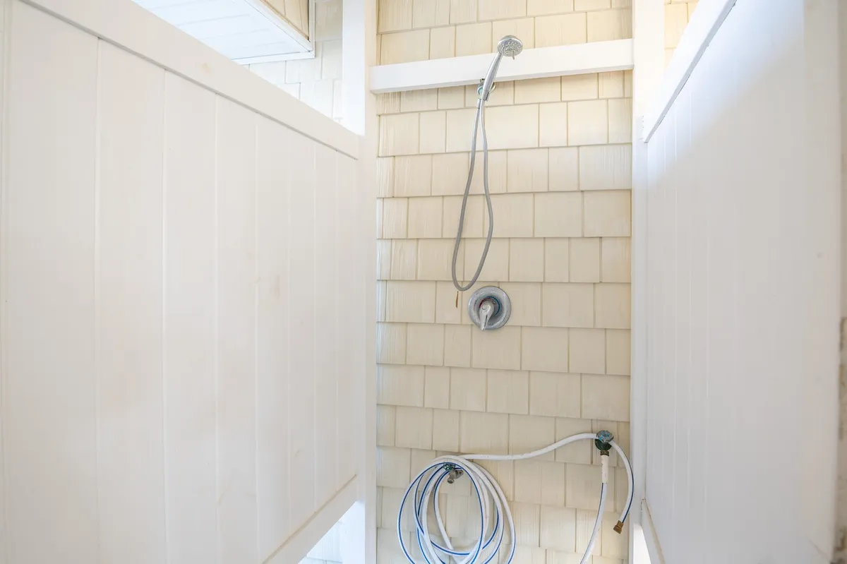 A clean, simple outdoor shower fixture is mounted on a textured shingle wall, framed by white privacy panels. The space is designed for post-beach refreshment, blending functionality with a touch of coastal charm.