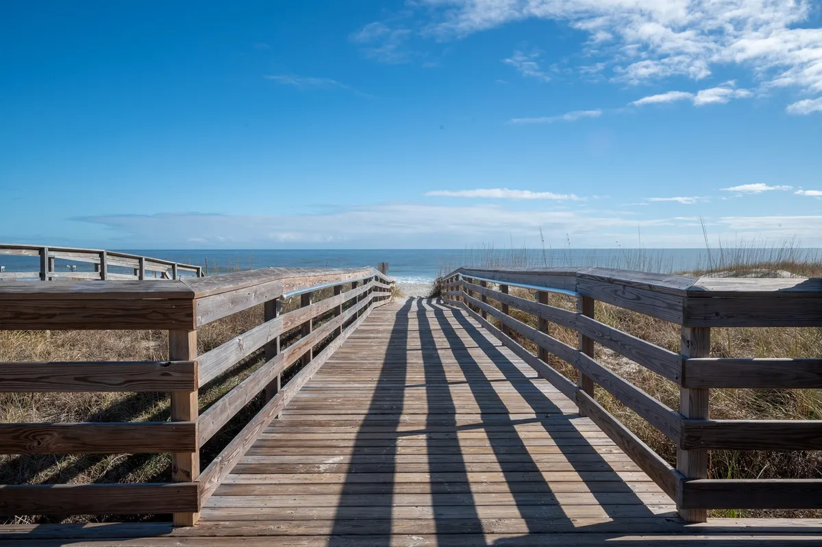 A stunning view of a wooden boardwalk leading directly to the ocean, with the blue sky and distant horizon providing a welcoming, serene backdrop. The vantage suggests easy access from the property to the beach, making it perfect for beach outings and sunset strolls.
