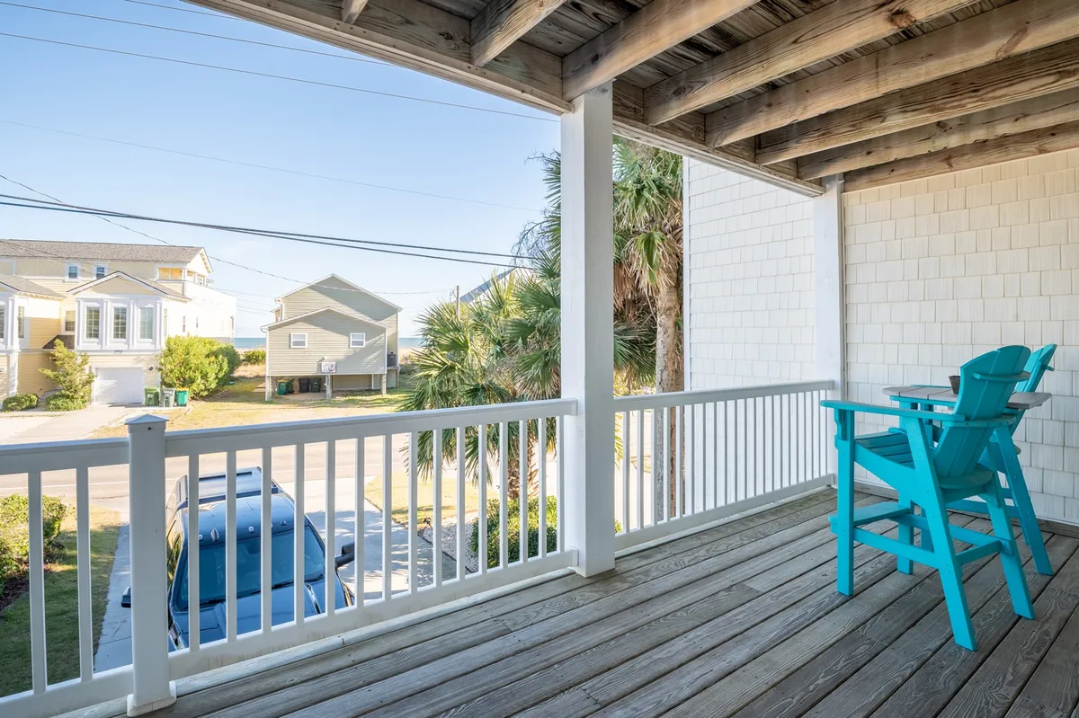 A wide view of the porch with blue Adirondack chairs facing outward, shaded by palm trees and overlooking a quiet street. The setup is perfect for relaxing and soaking in the ocean atmosphere, even from just one row back.