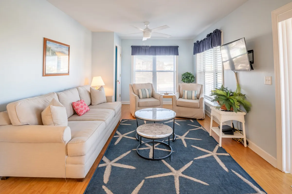 A sun-filled living room featuring a comfortable sofa and armchairs arranged around a coffee table, with a large window allowing natural light to pour in. Decorative touches, like a potted plant, add to the tranquil and inviting ambiance.