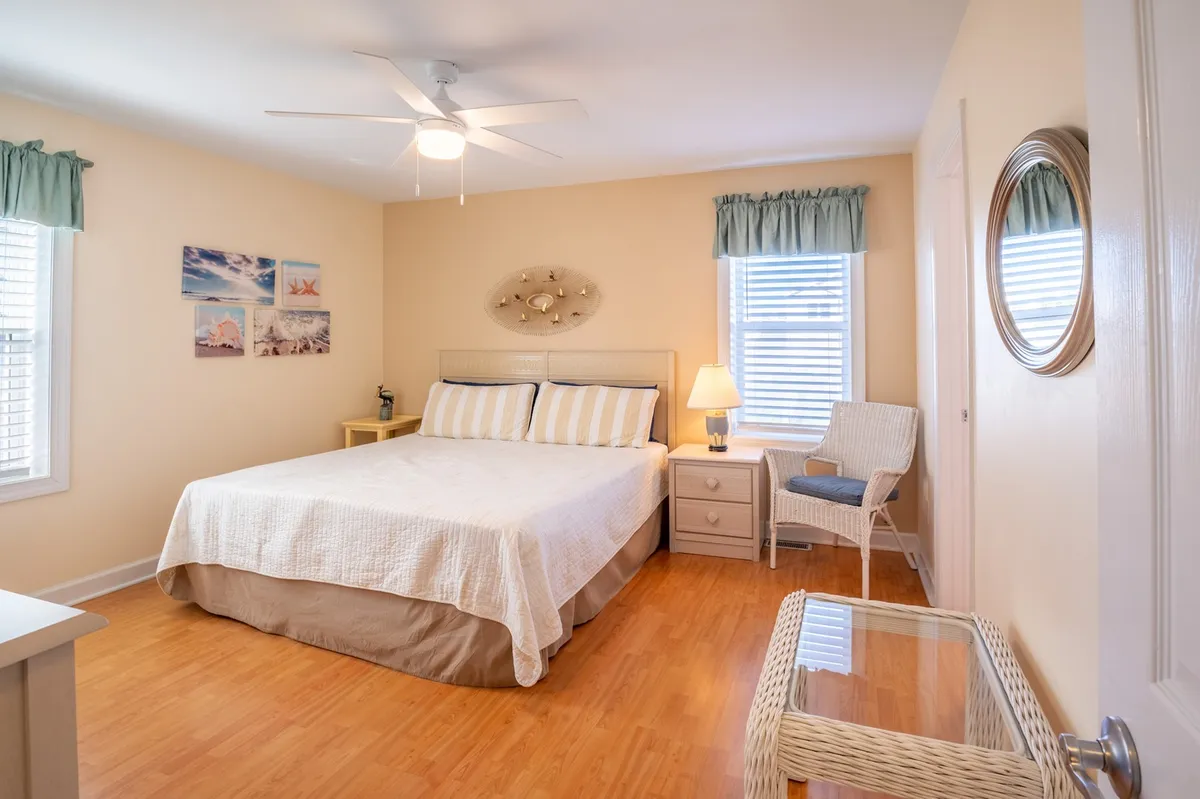 A serene bedroom styled with white bedding, a nightstand with a lamp, framed pictures on the wall, and a comfortable chair. The natural light filters in, enhancing the room's peaceful ambiance.