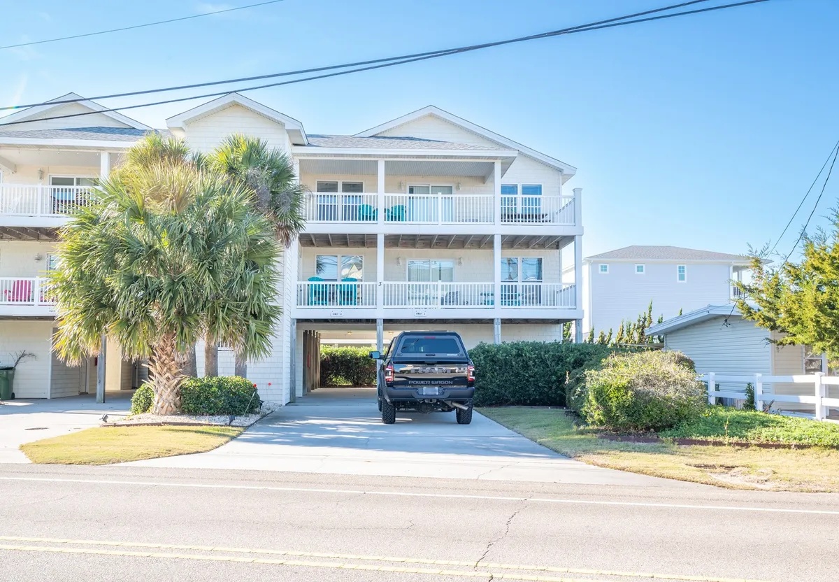 The image showcases the exterior of a welcoming condo just one row from the ocean, framed by a graceful palm tree. Spacious balconies with inviting blue chairs overlook a green yard, hinting at relaxing evenings watching sunsets or hosting family gatherings. A black truck parked nearby evokes the ease of arriving for a memorable beach getaway.