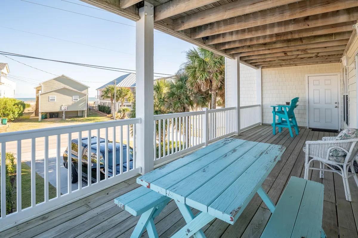 A bright exterior shot of the covered porch, featuring a blue picnic table surrounded by white railings and framed by palm trees. The scene exudes a casual, beachy vibe and highlights the inviting outdoor living space that's one row back from the ocean.