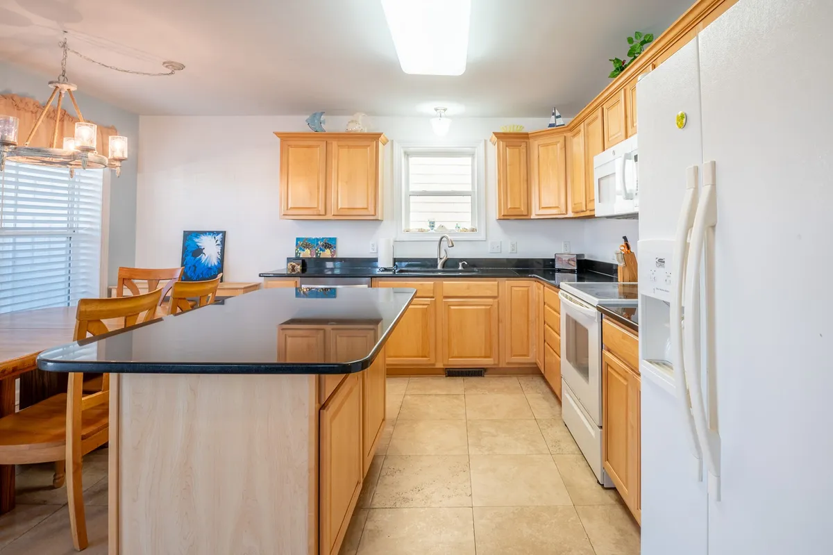 A view of the kitchen island with wooden cabinetry, black countertops, white appliances, and a glimpse of the adjacent dining area. The space is designed for easy meal prep, entertaining, and casual gatherings, with enough room for several people to move around comfortably.